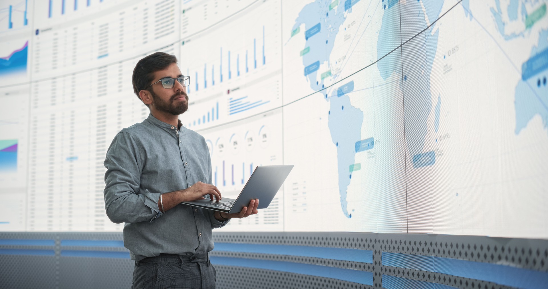 Indian Male Logistics Expert Holding Laptop Computer And Analyzing World Map On Big Digital Screen In Monitoring Office. Successful Man Developing New Efficient Routs For Global Product Distribution.